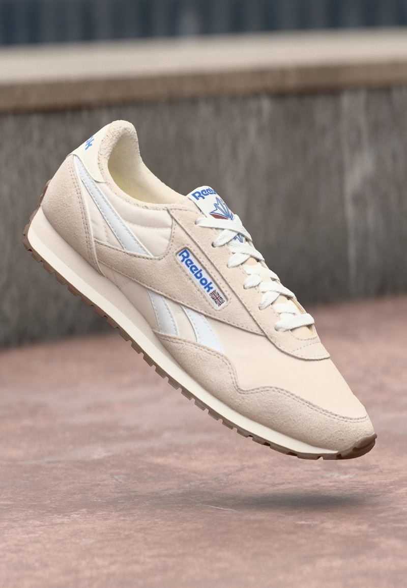 Beige zamšs un gluda ādas sporta apavi ar baltiem akcentiem, Reebok logo sānos un strukturēta zole, ar tradicionālām šņorēm.