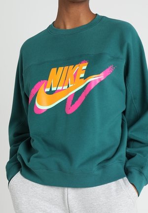 Person iført teal Nike sweatshirt med stort orange og gult Nike-logo og pink penselstrøg, kombineret med lysegrå bukser.