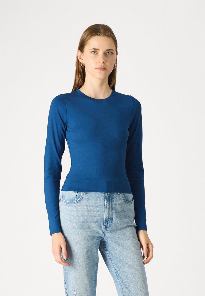 Μπλε τοπ μακρυμάνικο, εφαρμοστό, φτιαγμένο από απαλό ύφασμα; διαθέτει στρογγυλή λαιμόκοψη και ραφές χωρίς ένωση στους ώμους, σε συνδυασμό με ανοιχτόχρωμο μπλε τζιν.