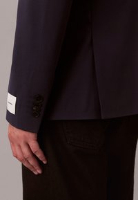 Hånd hvilende ved siden av ermet på en mørk marineblå blazer med fire svarte knapper og hvit Calvin Klein-label, båret over svarte bukser.