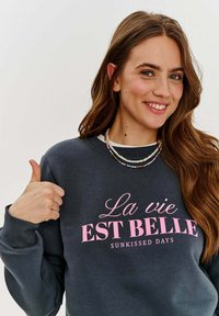 Uhlová mikina s ružovým nápisom "La vie EST BELLE SUNKISSED DAYS" a rebrovanými manžetami. Model má na sebe viacfarebný korálikový náhrdelník.