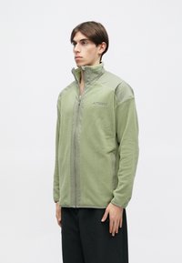 Bărbat tânăr îmbrăcat într-o jachetă din polar verde deschis cu fermoar, având logo-ul "Terrex", stând pe un fundal alb simplu și privind spre stânga.