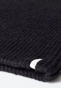 Zwart gebreid textiel met een geribbelde textuur, voorzien van een klein wit label aan de rand. Het oppervlak lijkt zacht en licht vezelig.