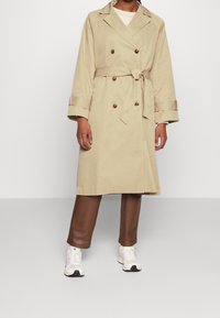 Beige trenchcoat met een dubbelgeknoopt ontwerp, tailleceintuur en knoopdetails. Gecombineerd met bruine broek en witte sneakers.