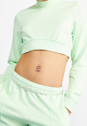 Close-up van een torso in een mentaalgroene cropped sweatshirt en bijpassende broek met een zilveren kettingriem en een navelpiercing met een lettercharme.