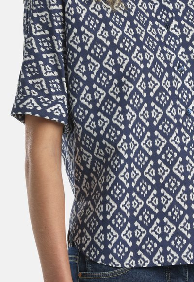 Tamsiai mėlyna trumpomis rankovėmis marškinėliai su baltu geometriniu raštu, užsegami sagomis priekyje, dėvimi su mėlynais džinsais, matomi nuo juosmens iki vidurio rankos.