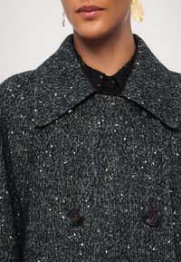 Personne portant un manteau texturé en chevrons gris foncé avec un grand col et des boutons noirs, associé à une chemise noire et des boucles d'oreilles dorées en forme de fleur.