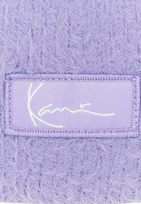 Paarse fuzzy stof met een rechthoekige stoffen patch met een witte handtekeninglogo. De textuur lijkt zacht en pluizig.