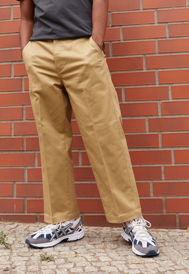 Pantaloni beige a gamba dritta con tasche laterali, indossati con scarpe da ginnastica grigie e blu, davanti a uno sfondo di muro di mattoni.