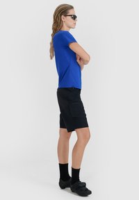 Personne aux cheveux longs portant une chemise bleue, un short noir, des chaussettes noires, des chaussures noires et des lunettes de soleil foncées, debout les bras croisés, de côté.