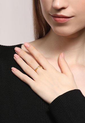 Gold- und silberner gedrehter Ring mit einer kleinen Reihe klarer Steine, getragen an einer Hand mit hellrosa Nägeln, vor einem dunklen Pullover-Hintergrund.
