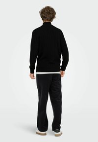 Mann mit lockigem Haar, der einen schwarzen, grob gestrickten Pullover, schwarze Hose und weiße Sneakers trägt, steht mit dem Rücken zu einem schlichten Hintergrund.