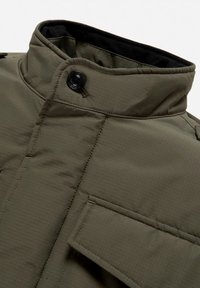 Colletto di giacca imbottita verde oliva con chiusura a bottone e tasca con pattina sul petto, caratterizzata da dettagli di cuciture trapuntate.