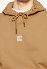 Sweat à capuche marron clair avec cordons noués en un nœud, comportant un petit patch logo crème centré de la marque The North Face.