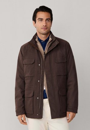 Chaqueta de tela marrón con un diseño estructurado, múltiples bolsillos frontales y un forro interior en contraste. Cuenta con cierre de cremallera y broches a presión.