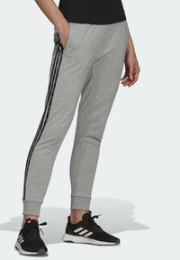 Grå sportbyxor med svarta sidosträck, avslutade vid vinklarna, och sidofickor, tillsammans med svarta sneakers med vita accenter.