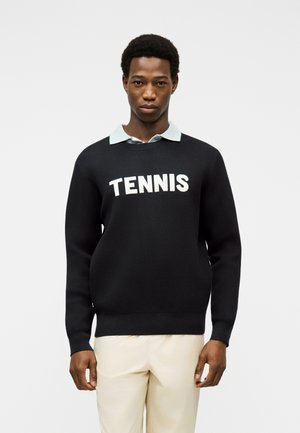 Tânăr purtând un pulover negru cu inscripția „TENNIS”, o cămașă albastru deschis cu nasturi la guler și pantaloni albi, stând în picioare pe un fundal simplu.