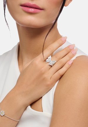 Woman wearing a silver butterfly-shaped ring and a delicate flower bracelet, showing manicured nails and smooth skin on arm and shoulder.