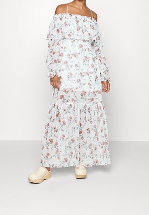 Vrouw draagt een lange witte off-shoulder jurk met bloemenprint, volants en doorschijnende lange mouwen, gecombineerd met crèmekleurige houten klompen.