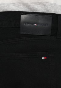 Vaqueros de mezclilla negra con una etiqueta de cuero de la marca "TOMMY HILFIGER". Presentan un costura del logo en contraste en rojo, blanco y azul en el bolsillo.