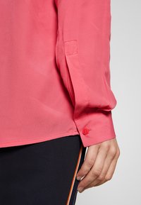 Lightweight, semi-sheer, coral button-up shirt with long sleeves, featuring a single red button at the cuff and a smooth texture.