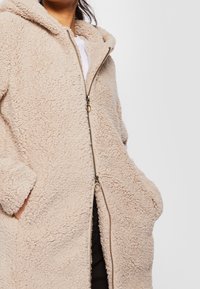 Cappotto in pile beige con cappuccio, dotato di zip frontale, ampie tasche e superficie morbida e testurizzata. I polsini sono leggermente arrotolati.
