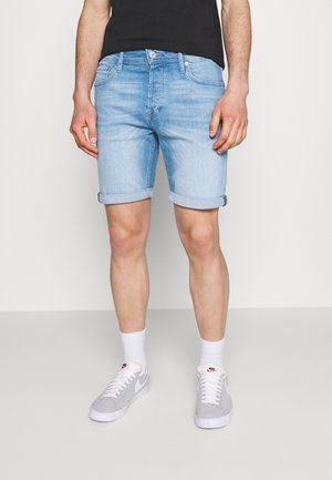 Mężczyzna ubrany w jasnoniebieskie podwinięte szorty jeansowe, białe skarpetki, szare buty Nike i czarną koszulkę, stoi na jednolitym tle.