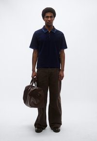 Μπλε ναυτικό πόλο πουκάμισο με αντίθετο γιακά, καφέ παντελόνι με φαρδιά πόδια και καφέ δερμάτινη σακούλα τύπου duffel. Λείο υφή, μοντέρνος σχεδιασμός.