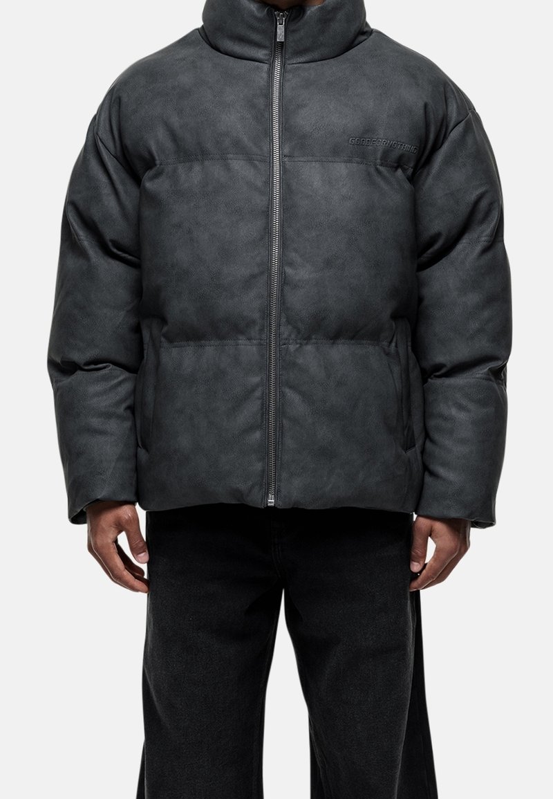 Homme portant une veste matelassée noire à fermeture éclair avec col montant et poches latérales, associée à un pantalon noir sur un fond uni.