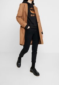 Brun kappa över svart hoodie med metalliska grafiker, tillsammans med svarta skinny jeans och svarta skor. En enkel och modern design.