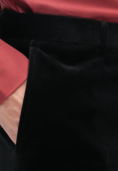 Černé sametové kalhoty s měkkou texturou a boční kapsou. Částečně viditelná je rustikálně zbarvená košile s hladkým materiálem a manžetou.