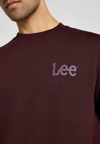 Uomo che indossa una felpa girocollo di un rosso scuro con un logo "Lee" di color lilla sul petto.