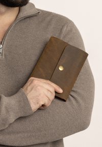 Bruine leren portemonnee met een drukknoopsluiting en een gladde textuur, vastgehouden tegen een grijze trui-arm. Netjes, rechthoekig ontwerp.