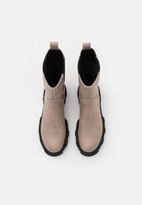 Ett par beige ankelskor med svarta elastiska sidor och tjocka svarta sulor, sedd uppifrån mot en vit bakgrund.