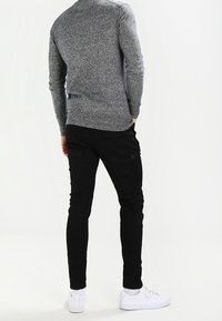 Harmaan tekstuuriseen neuleeseen, mustiin tiukkoihin housuihin ja valkoisiin tennareihin pukeutunut henkilö näkyy selkäpuolelta. Asusteessa on yksinkertainen ja siisti muotoilu.