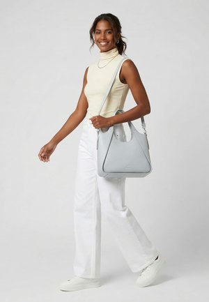 Femme souriante, marchant, portant un haut beige sans manches, un pantalon blanc, des baskets blanches, et un sac à main gris clair sur l'épaule, devant un fond uni.