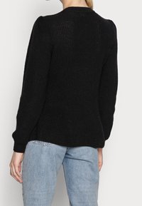Schwarzer Strickpullover mit langen Ärmeln, gerippter Textur und Rundhalsausschnitt. Der Rücken hat einen geraden Schnitt und eine lockere Passform. Kombiniert mit hellblauen Jeans.