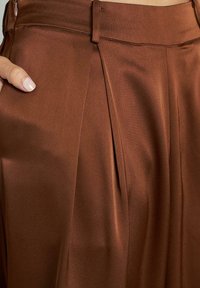 Brązowe, plisowane spodnie wykonane z gładkiego materiału, z dopasowanym paskiem w talii i detalami w formie bocznych kieszeni. Faktura wydaje się miękka i lekko błyszcząca.