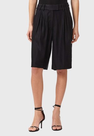 Vrouw die zwarte, hooggetailleerde, wijd uitlopende shorts tot over de knie draagt en zwarte, bandjes-achtige hoge hakken met hakken, staand tegen een effen achtergrond.