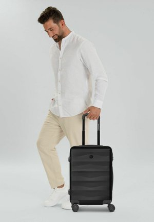 Hombre vestido con camisa blanca y pantalones beige tirando de una maleta rígida negra con asa extendida y cuatro ruedas.