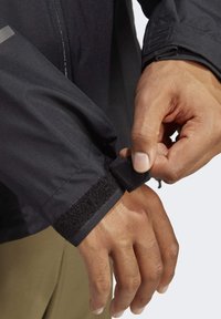Giacca nera impermeabile con polsini regolabili in Velcro. Primo piano di una mano che sistema la cinghia, evidenziando la texture liscia e il design moderno.