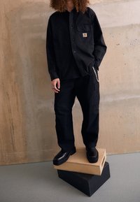 Musta napillinen paita, jossa on rintatasku, yhdistettynä löysiin housuihin ja tukevat mustat kengät, jotka seisovat pinottujen laatikoiden päällä.