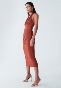 Rust-colored, mid-length dress with halter neckline and gathered detailing. Smooth fabric with a fitted silhouette and side ruching.