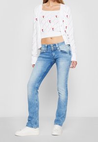 Ljust blå denimjeans med rak bendesign, utrustade med två knappar, i kombination med en vit, stickad cardigan och en kort topp.