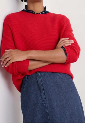 Personne les bras croisés portant un pull côtelé rouge, une chemise à col foncé à motifs et une jupe en jean bleue.