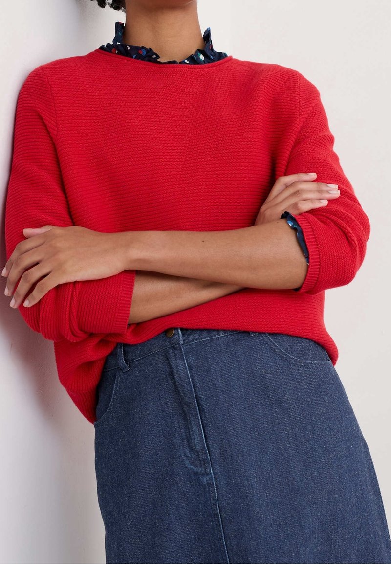 Personne les bras croisés portant un pull côtelé rouge, une chemise à col foncé à motifs et une jupe en jean bleue.