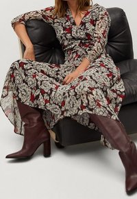 Sukienka maxi z kwiatowym wzorem w kremowym, czerwonym i czarnym; długie rękawy i dekolt w kształcie litery V. Połączona z burgundowymi botkami na słupku do kolan na sofie.