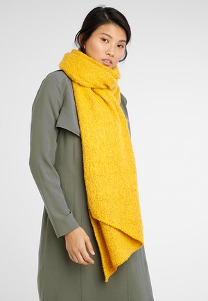 Écharpe texturée jaune vif en matériau doux, drapée autour du cou, avec une forme lâche et décontractée, portée sur un vêtement gris.