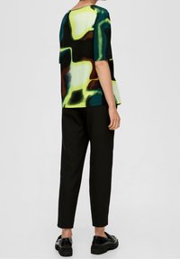 Bluzka z krótkim rękawem w abstrakcyjny wzór w odcieniach zieleni, czerni i brązu, zestawiona z czarnymi, zwężanymi spodniami i czarnymi butami.