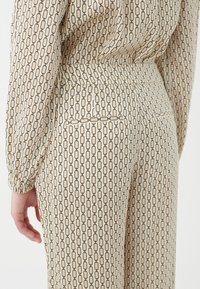 Tissu crème avec un motif de maillons de chaîne noirs récurrents. Le design comprend une poche arrière et des manches à revers.